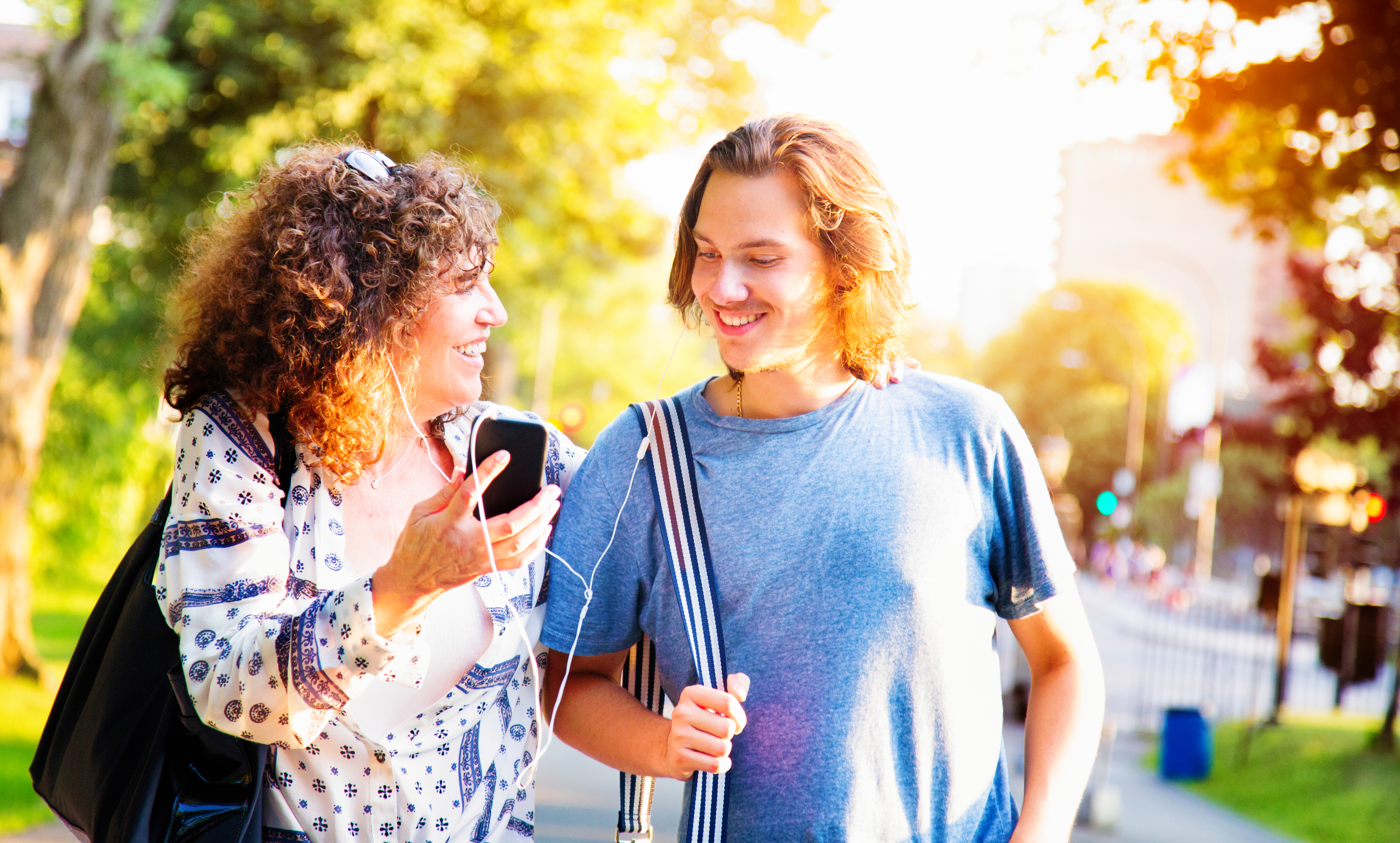 Smiling woman with curly hair holding smartphone and wearing earbuds, with a man's arm around her shoulder outdoors